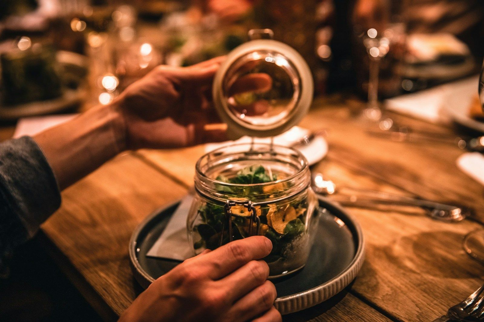 Eine Person hebt den Deckel eines Glasbehälters und zeigt einen frischen Salat auf einem Holztisch mit verschiedenen Geric...