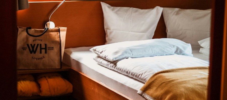Gemütliches Schlafzimmer mit zwei weißen Kissen, einer braunen Decke und einer Tasche neben einer Nachttischlampe.