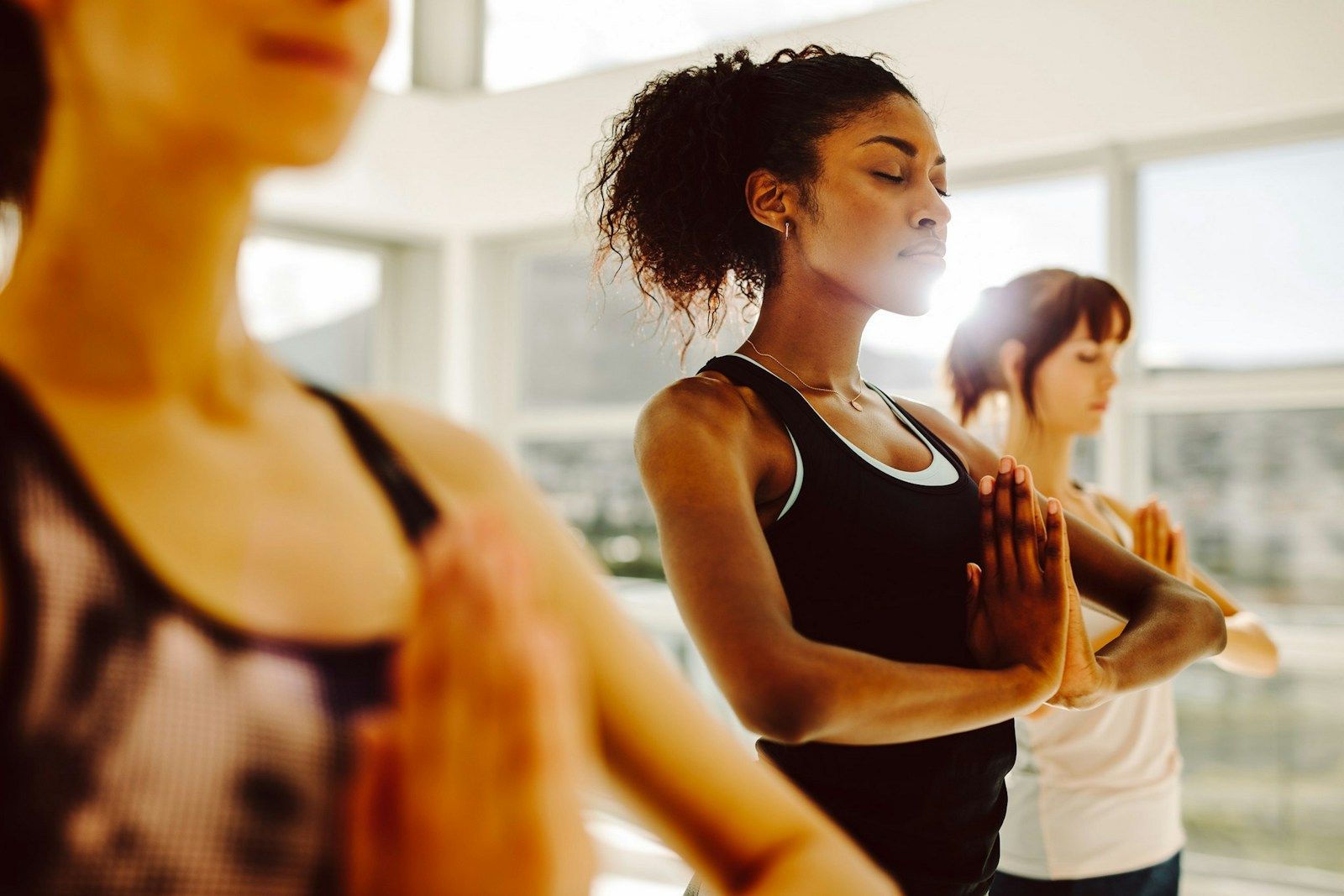 Drei Frauen praktizieren Yoga drinnen, Hände in Gebetsposition, mit Sonnenlicht, das hereinströmt.