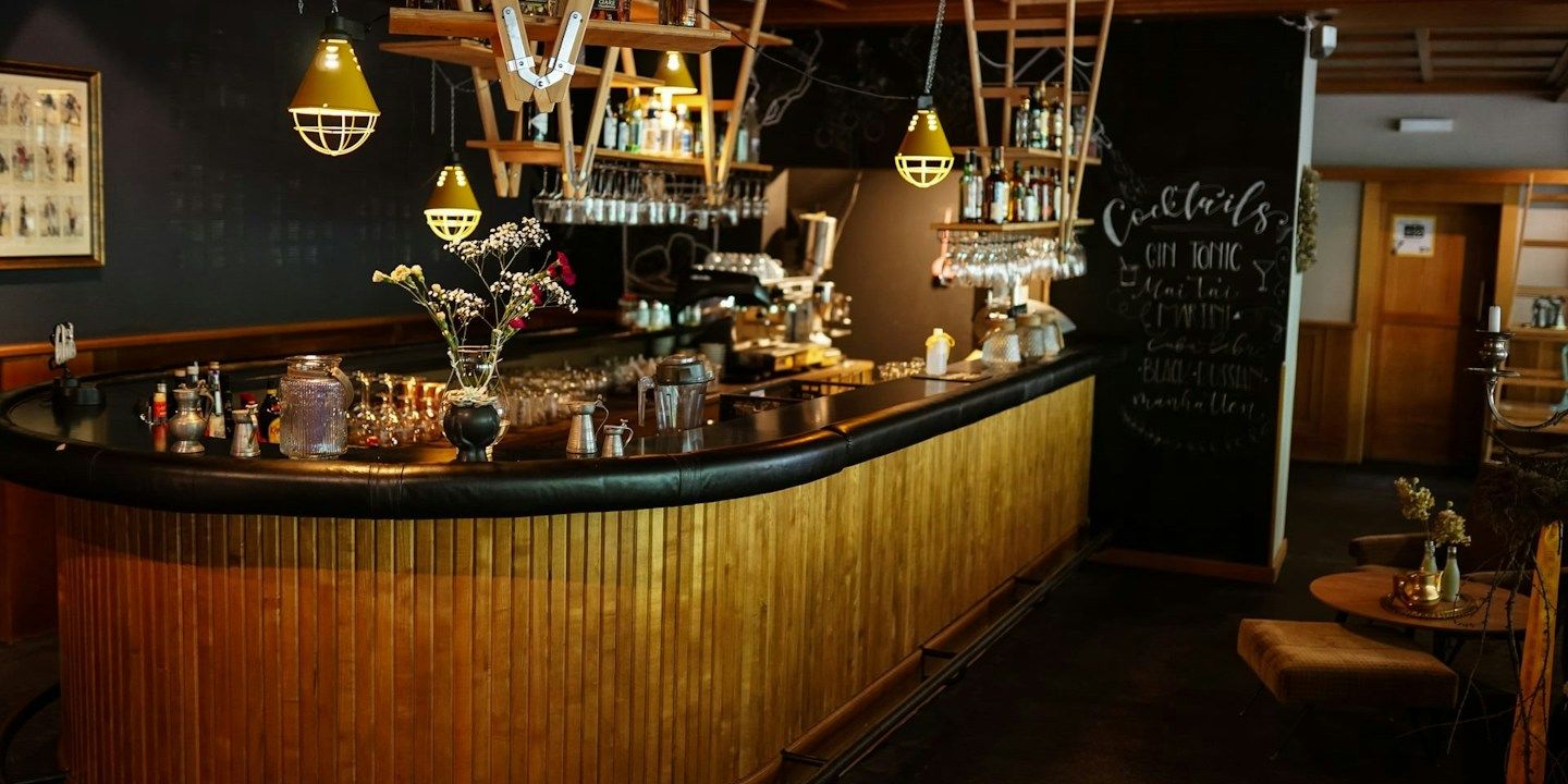 Eine stilvolle Bar mit einer Holztheke, ausgestellten Flaschen und Hängeleuchten, dazu eine Tafel mit Getränken.