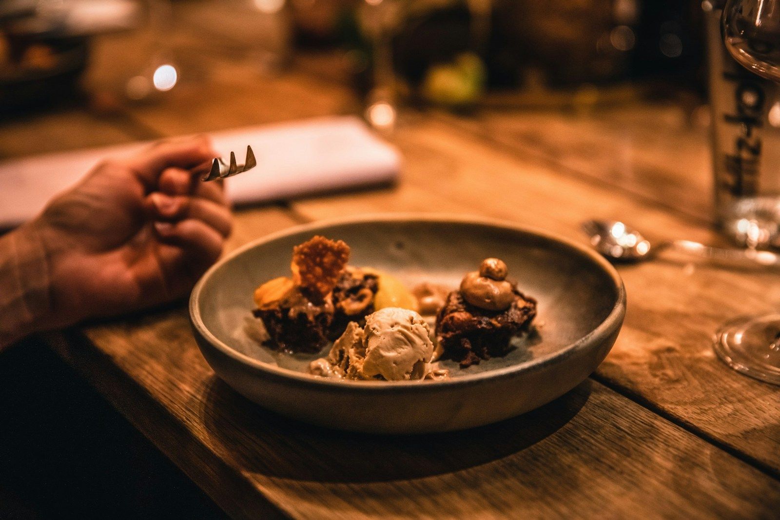 Eine Hand mit einer Gabel neben einem Teller mit Desserts und Eiscreme auf einem Holztisch.