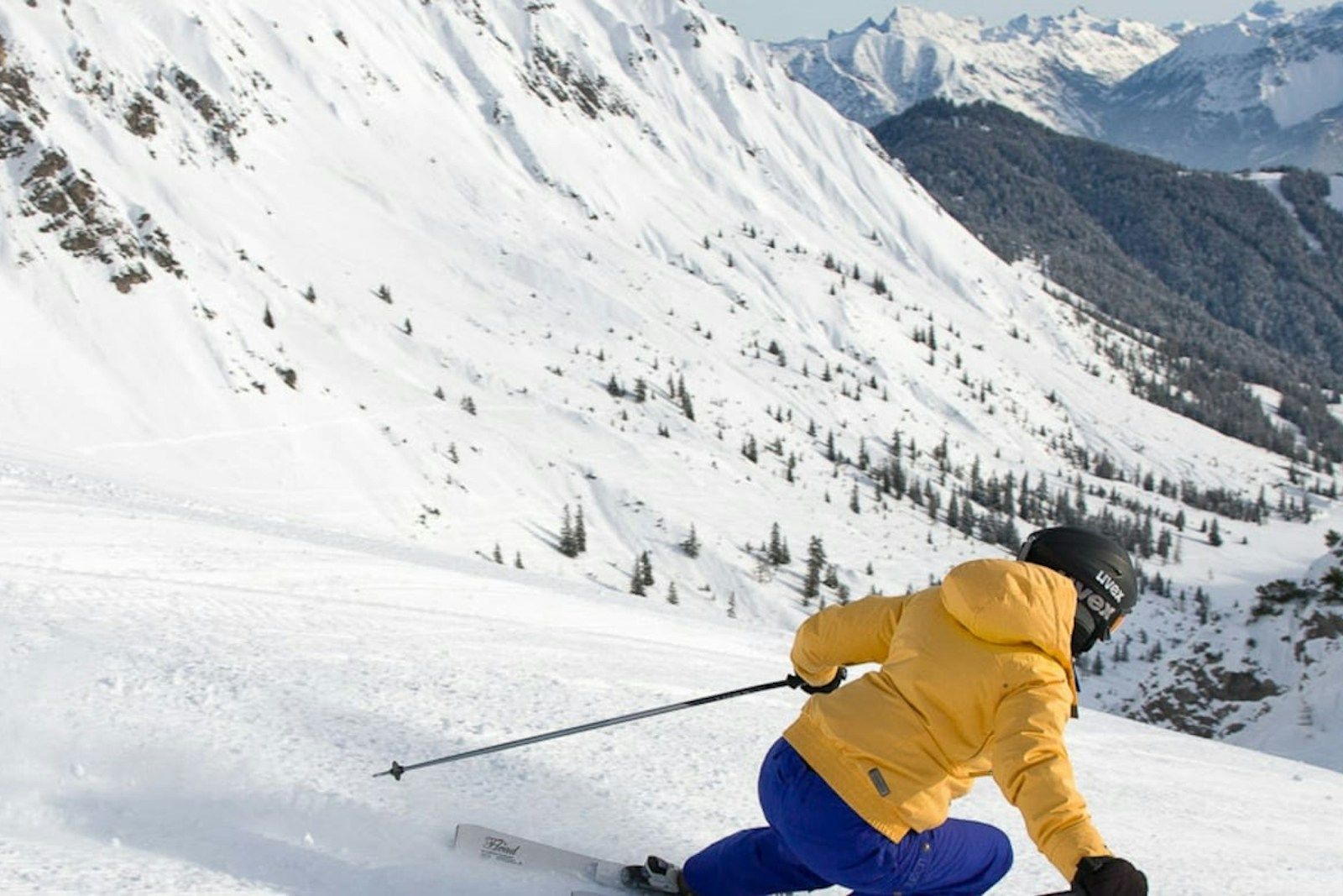 Ein Skifahrer in einer gelben Jacke fährt einen verschneiten Hang, umgeben von Bergen.