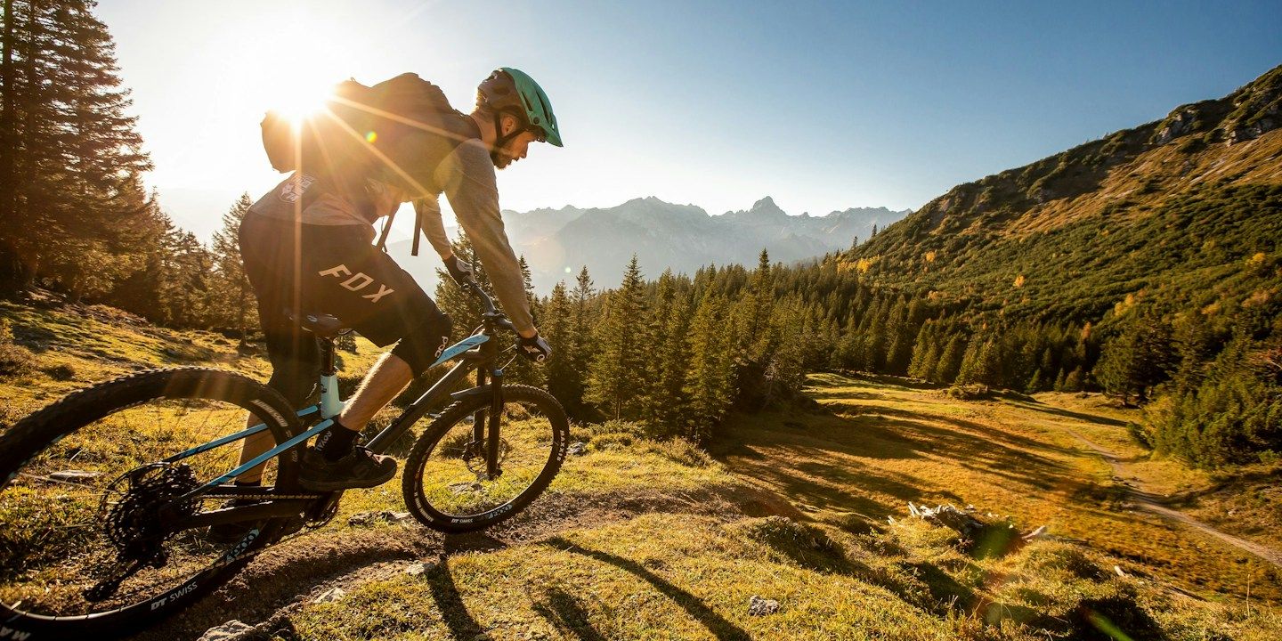 Ein Mountainbiker fährt auf einem Pfad mit der Sonne im Rücken, umgeben von Bäumen und Bergen.