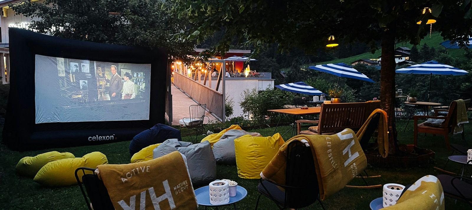 Außenkino mit großer Leinwand, Sitzsäcken und gemütlichen Sitzplätzen unter Bäumen in der Dämmerung.