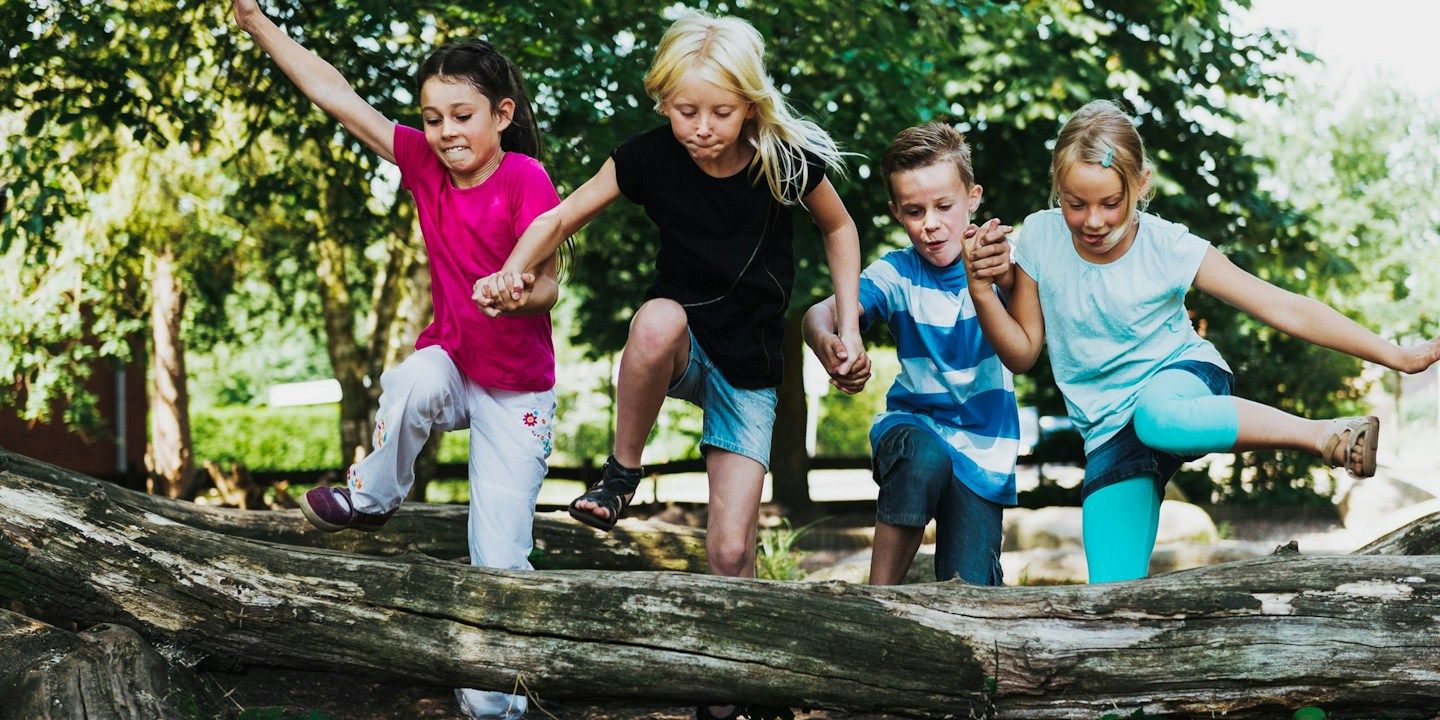 Vier Kinder springen fröhlich über einen Baumstamm in einer sonnigen Umgebung.