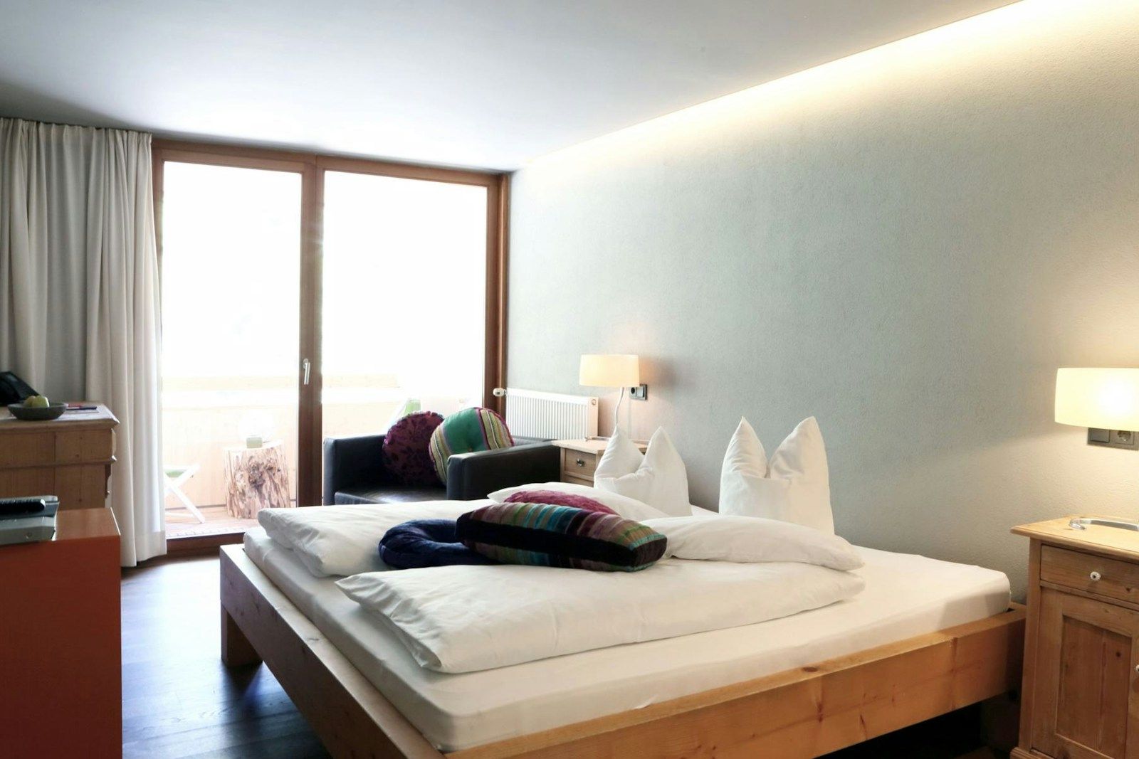 Helles Hotelzimmer mit großem Bett, Holzmöbeln und einem Balkonbereich mit Sofas.