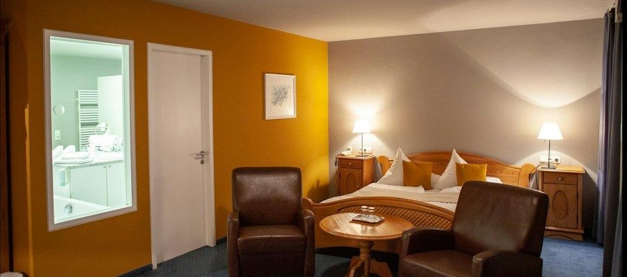Ein gemütliches Hotelzimmer mit einem Bett, zwei Sessel und einem kleinen Tisch; gelbe und graue Wände.