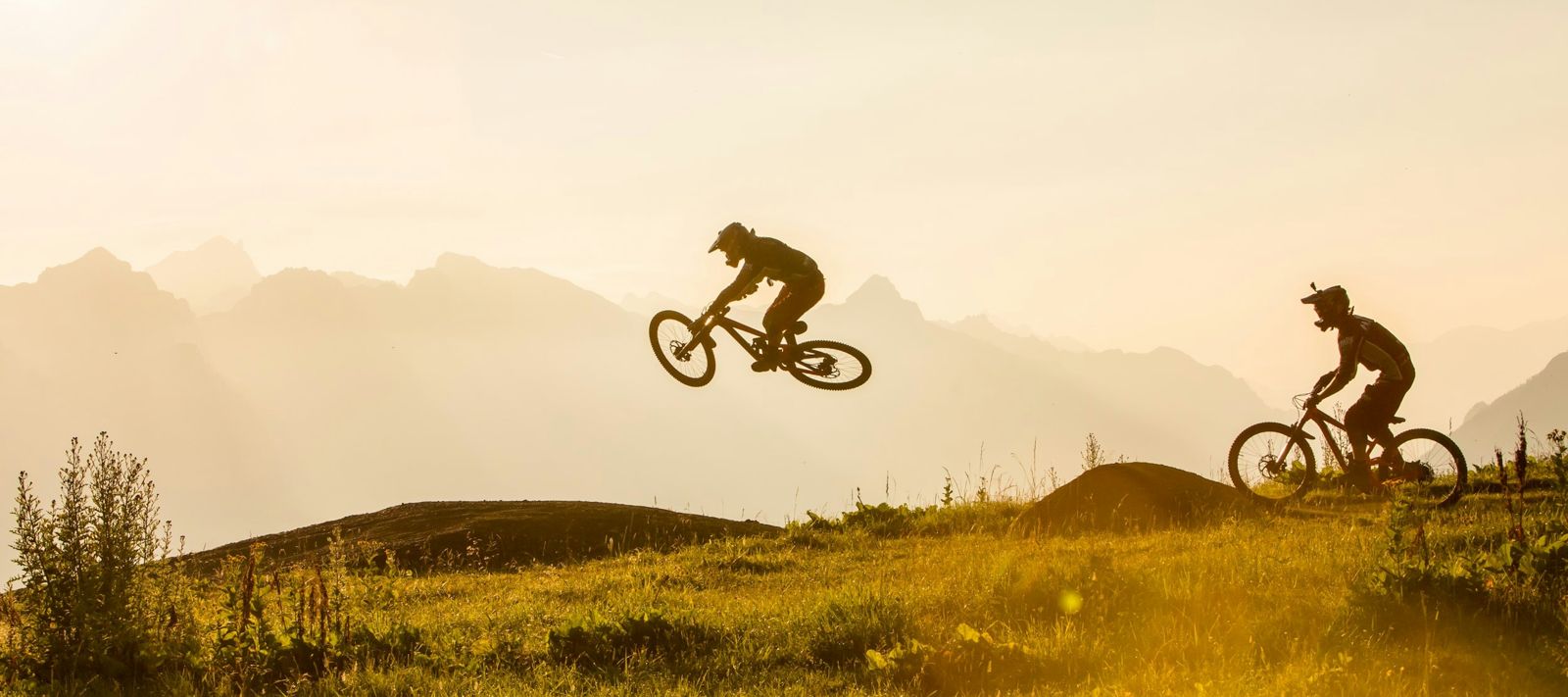 Zwei Mountainbiker springen über einen Hügel bei Sonnenuntergang, mit Bergen im Hintergrund.