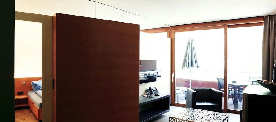 Innenansicht eines modernen Hotelzimmers mit einer Holztrennwand, großem Fenster und Sitzgelegenheiten auf dem Balkon.