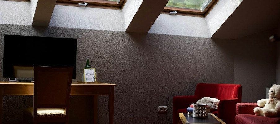 Ein gemütlicher Dachraum mit Dachfenstern, einem Schreibtisch, einem Fernseher, roten Stühlen und einem Teddybären auf ein...