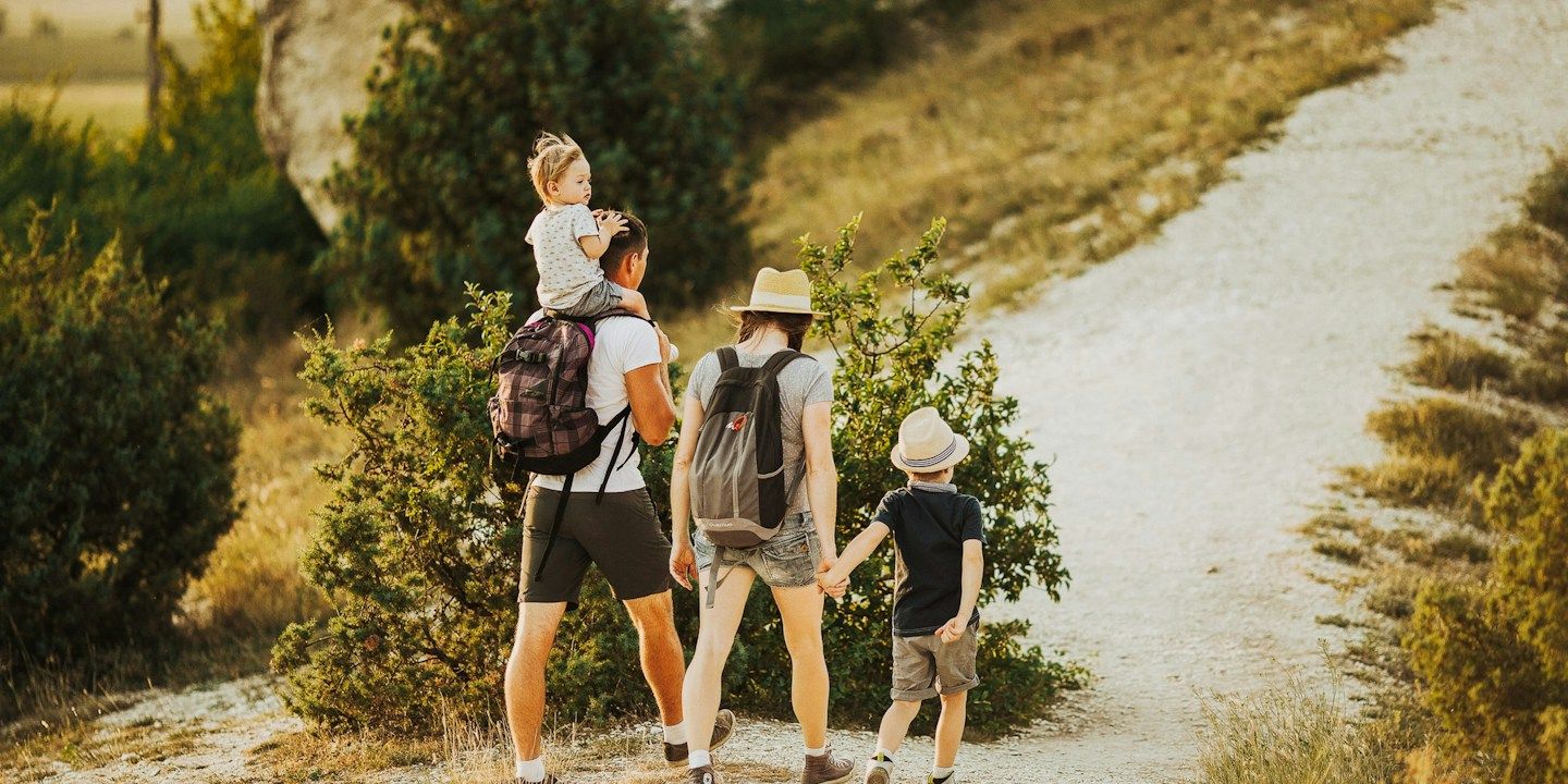 Eine Familie wandert auf einem Pfad; ein Erwachsener trägt ein Kind, während ein anderer Erwachsener und ein Junge Händche...