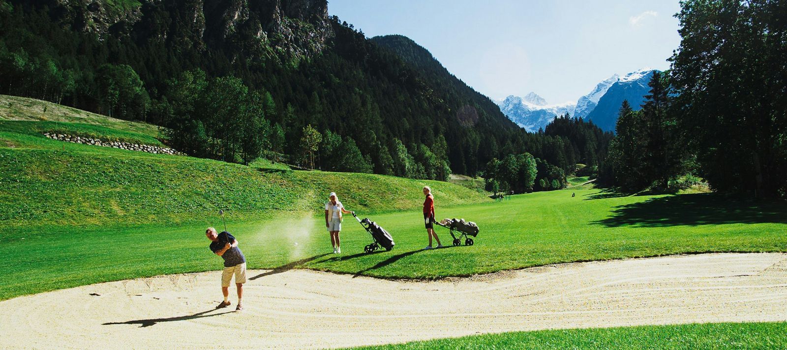 Drei Golfer auf einem sonnigen Platz; einer ist im Bunker, während die anderen mit ihren Schlägern zuschauen.