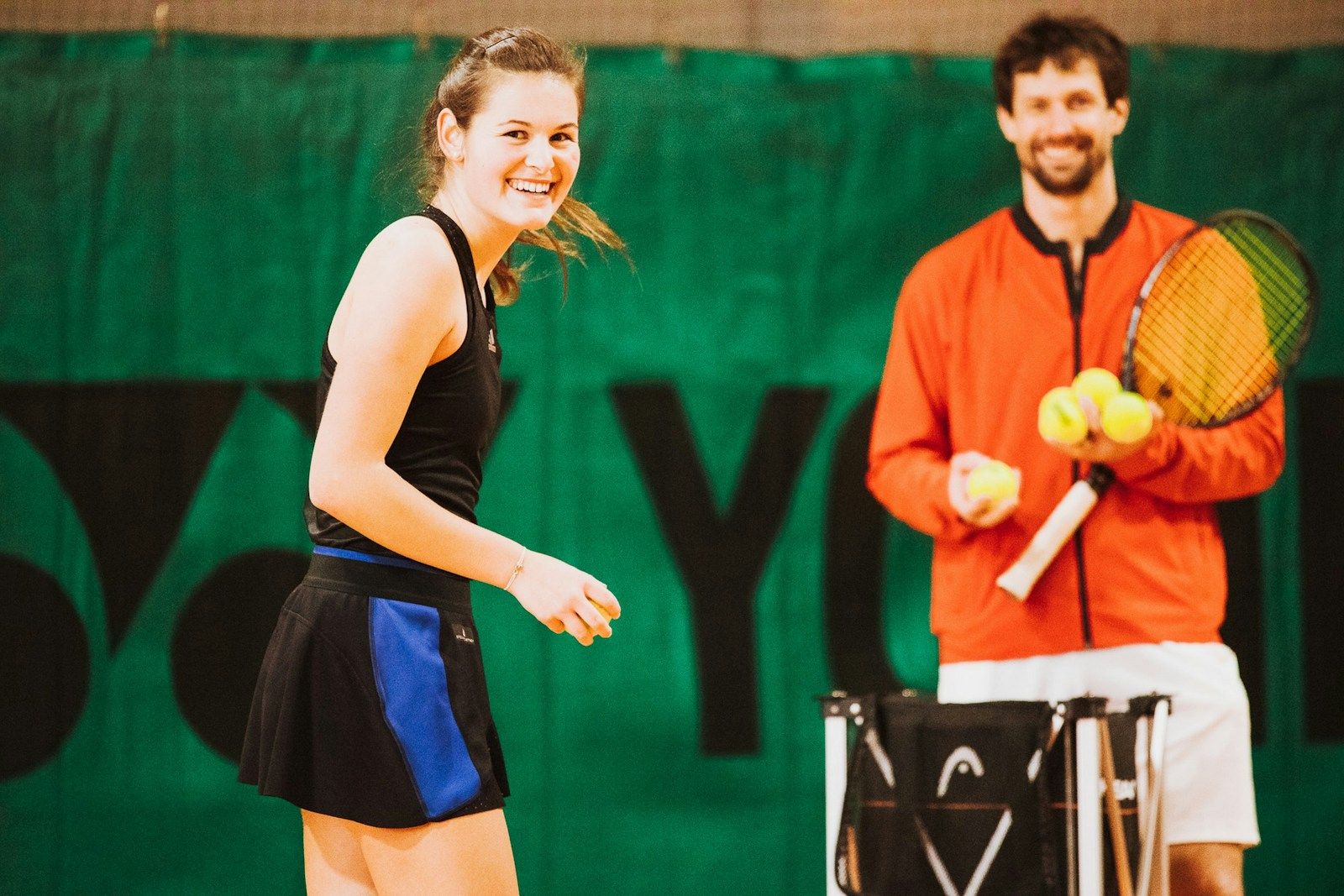 Eine lächelnde junge Frau in einem Tennisoutfit steht auf dem Platz, während ein Mann in einer roten Jacke Tennisbälle häl...