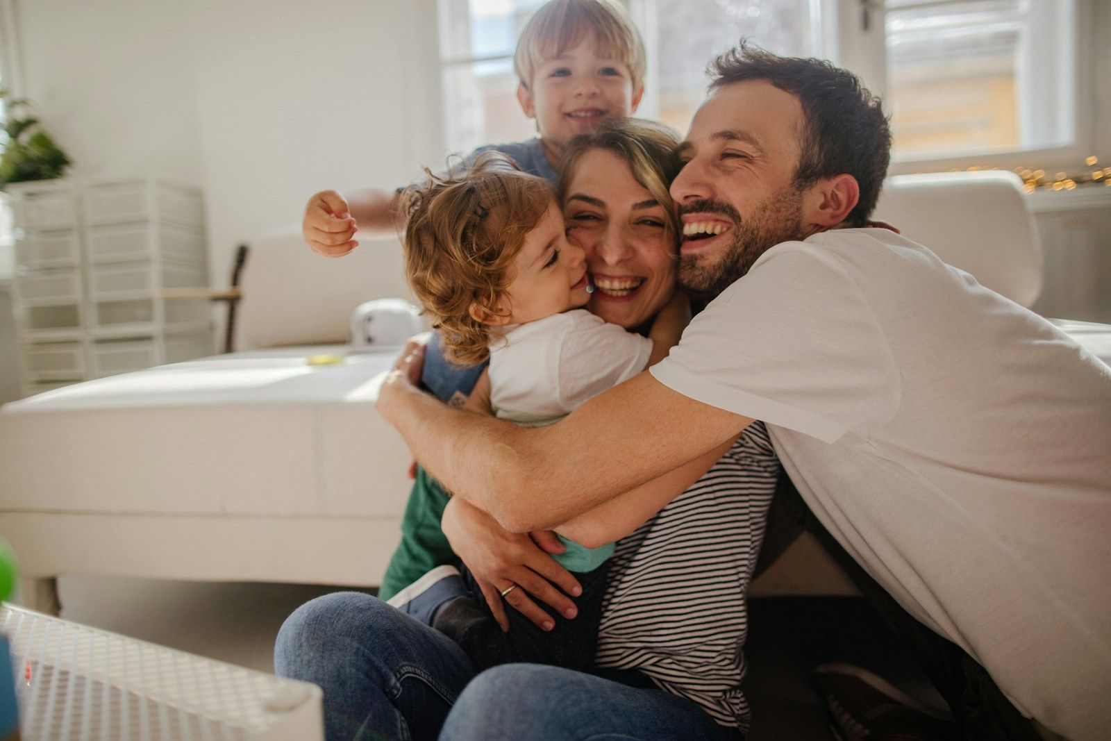 Eine Familie von vier Personen umarmt sich fröhlich in einem hellen Wohnzimmer und genießt die gemeinsame Zeit.