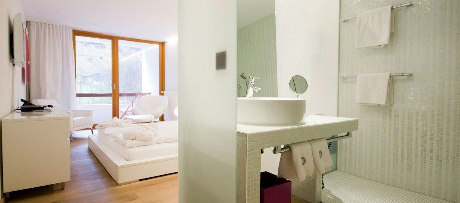 Modernes Hotelzimmer mit Bett, weißer Einrichtung und einem offenen Badezimmerbereich mit Waschbecken und Dusche.