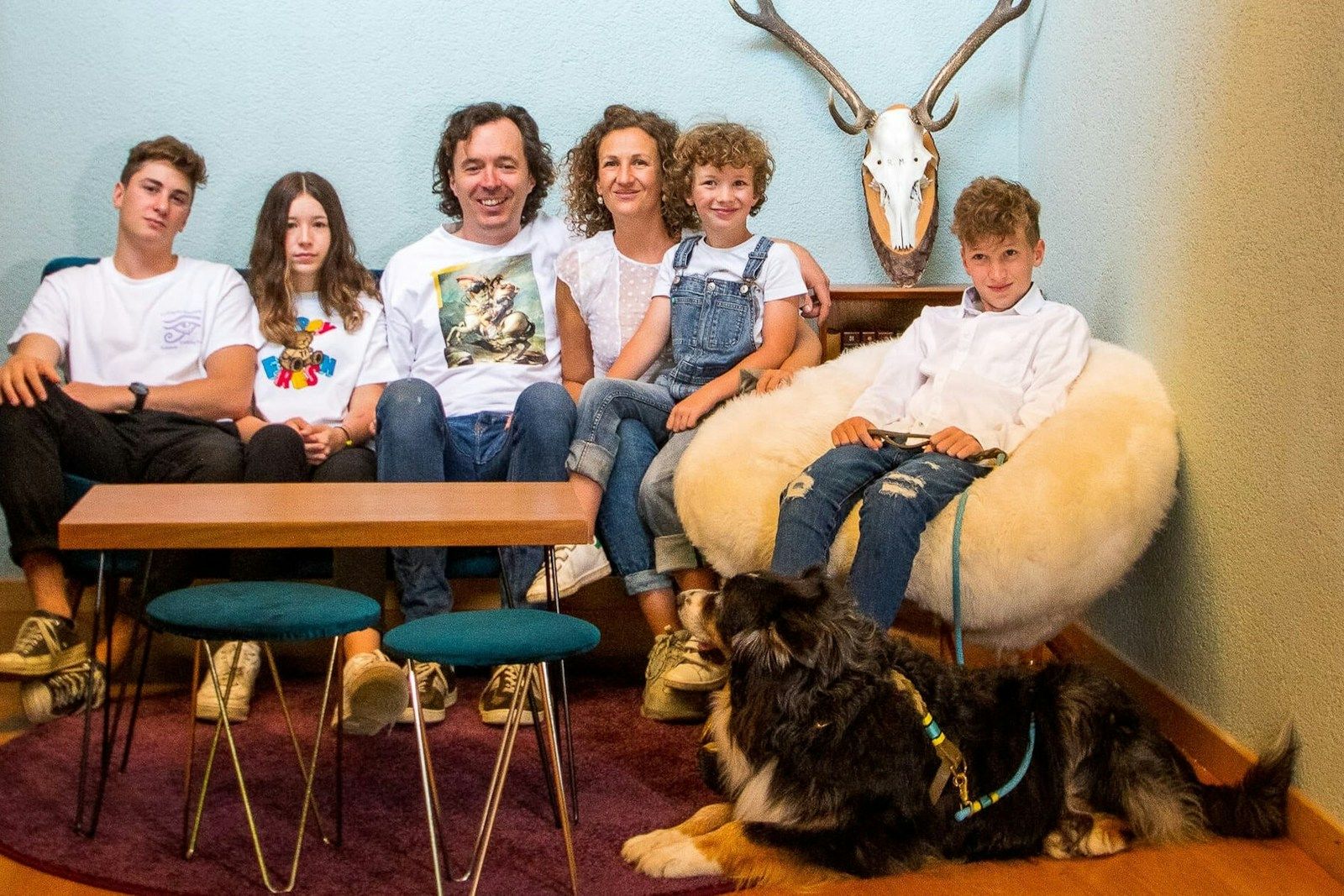 Eine fünfköpfige Familie und ein Hund sitzen in einem gemütlichen Wohnzimmer mit einem Holztisch und dekorativer Wand.