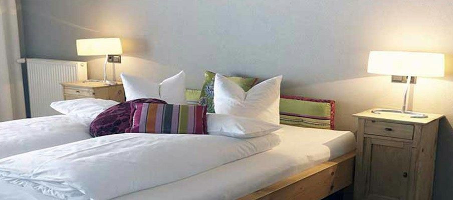 Ein gemütliches Schlafzimmer mit zwei Betten, hellen Lampen und bunten Kissen auf einem Holzrahmen.