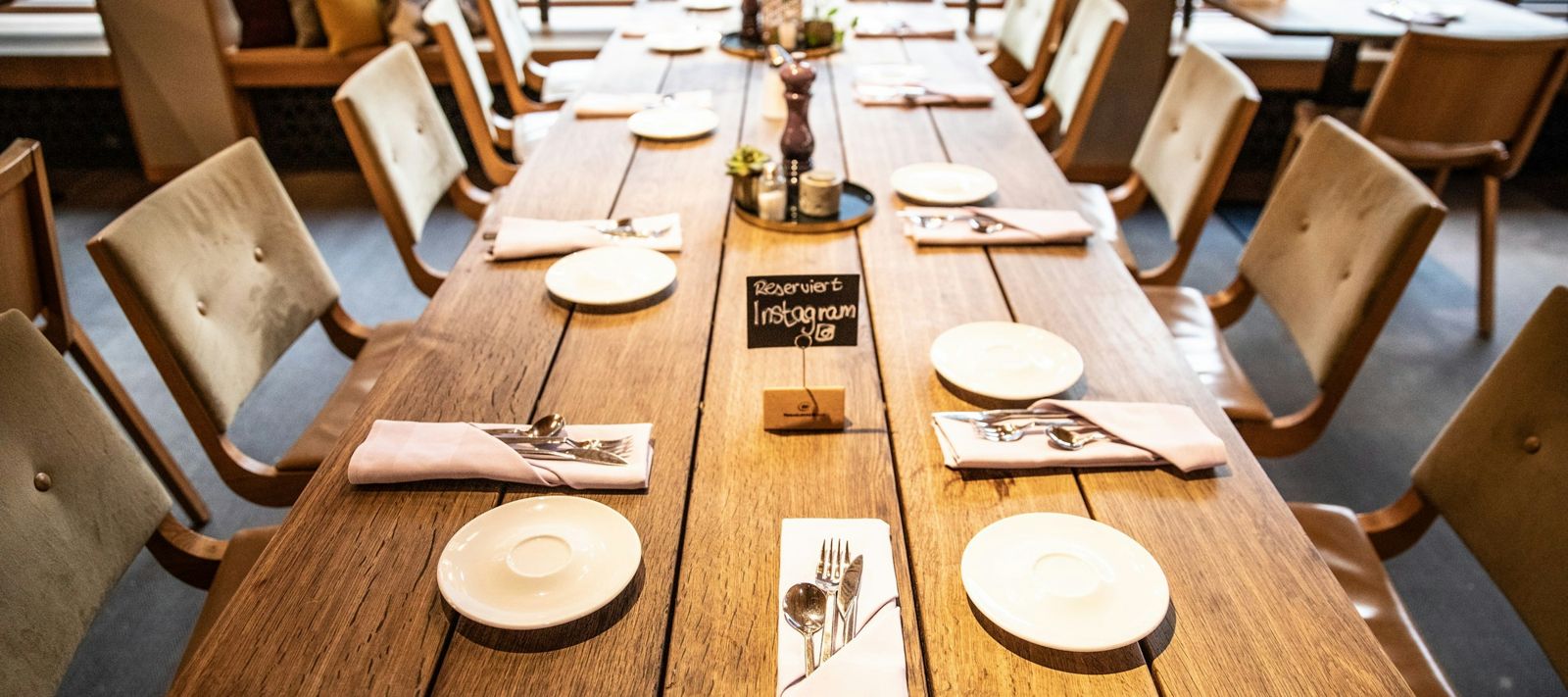 Ein langer Holztisch im Restaurant, gedeckt mit Tellern, Besteck und Servietten, umgeben von Stühlen.