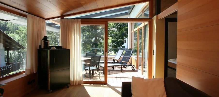 Gemütlicher Raum mit Holzwänden, einem Sofa und großen Fenstern, die zu einer Terrasse mit Liegestühlen führen.