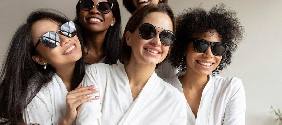 Fünf Frauen in weißen Bademänteln und Sonnenbrillen lächeln in einem hellen Raum zusammen.